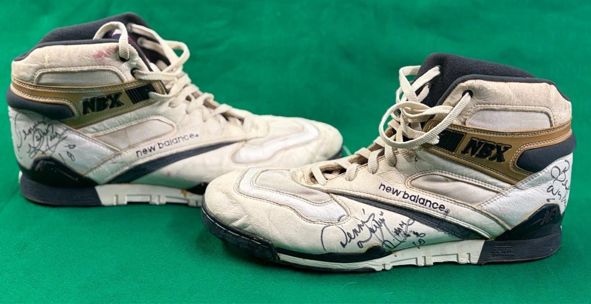 Pair of white, gold and navy New Balance NBX shoes signed by Dennis Rodman on a green background